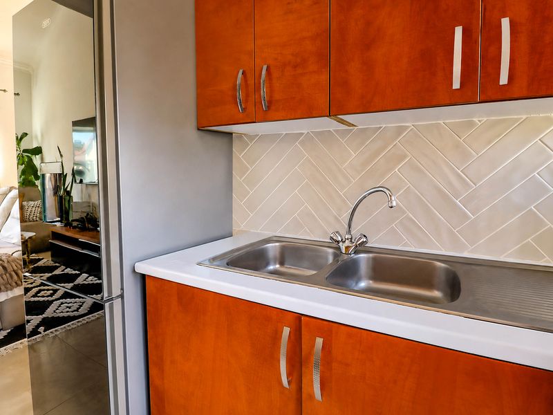 A compact kitchen area featuring warm-toned wooden cabinetry, a modern herringbone tile splashback, and a functional stainless steel double sink with a chrome mixer tap.