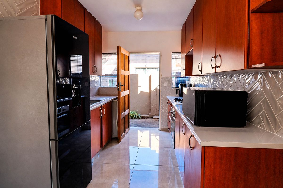 A functional galley-style kitchen featuring warm-toned cabinetry, a modern tiled backsplash, polished floor tiles, and direct access to an outdoor area.
