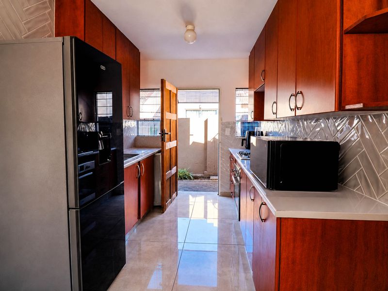 A functional galley-style kitchen featuring warm-toned cabinetry, a modern tiled backsplash, polished floor tiles, and direct access to an outdoor area.