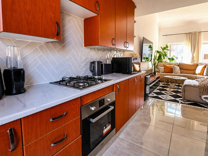 Modern open-plan kitchen featuring warm wood-toned cabinetry, a stylish herringbone backsplash, stone countertops, and integrated stainless steel appliances, flowing into the living area.
