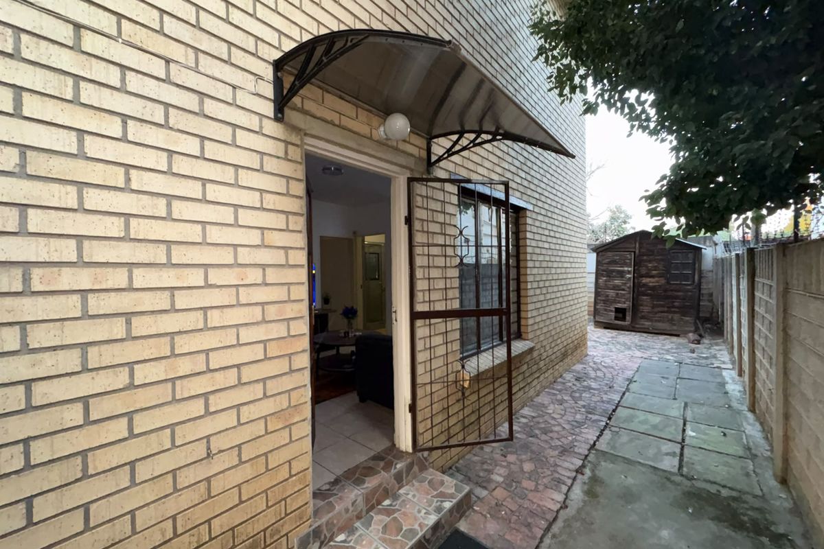Rear exterior view of the townhouse featuring a brick facade, a protective door awning, security gate, and a paved pathway leading to a garden shed.