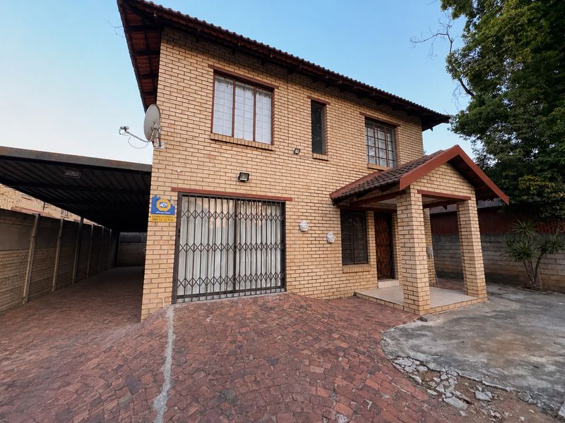Front elevation of a double-story face brick townhouse featuring a paved driveway, covered carport, and a covered entrance porch.