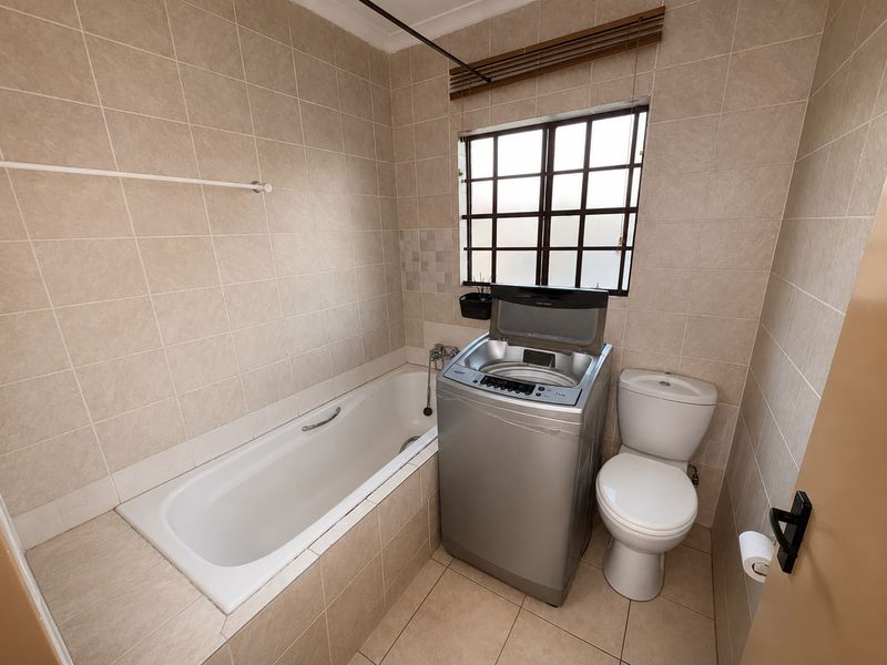 A functional bathroom featuring a built-in bathtub, toilet, and tiled finishes, with space currently utilized for laundry equipment.