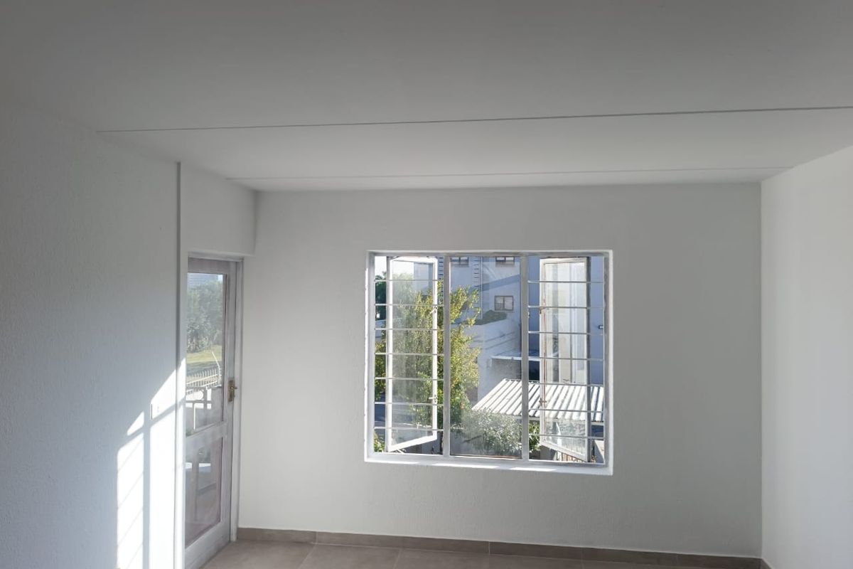 A bright, empty bedroom featuring neutral tiled flooring, a glass panel door, and windows fitted with security bars.