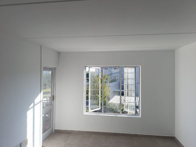 A bright, empty bedroom featuring neutral tiled flooring, a glass panel door, and windows fitted with security bars.