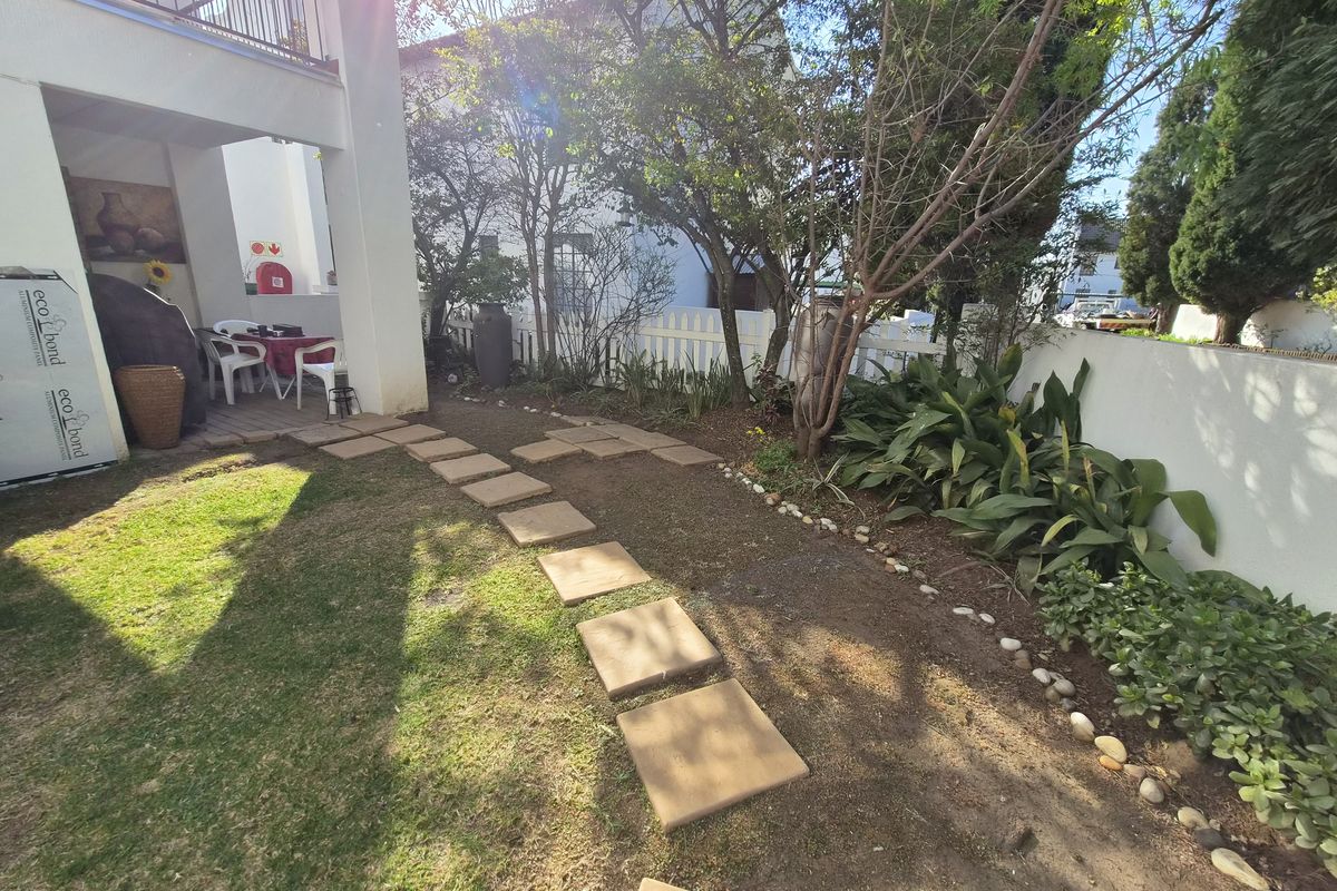 A private rear garden featuring a covered patio area, paved stepping stones, and established landscaping enclosed by a white boundary wall.