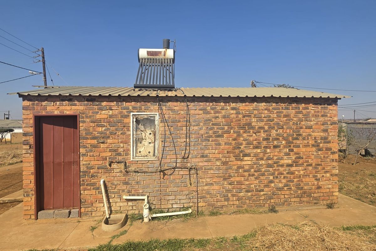 Rear view of a brick structure featuring a solar water heater, corrugated metal roof, and external plumbing connections.