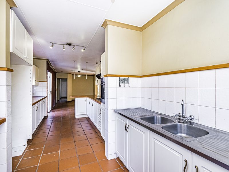 Spacious kitchen featuring built-in cabinetry, double basin sink, tiled splashbacks, and terracotta flooring, offering a functional layout with potential for modern updates.