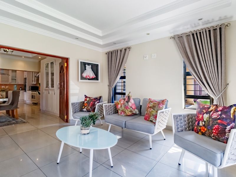 A bright and well-maintained living room featuring tiled flooring, decorative ceiling cornicing, and a seamless transition into the adjacent kitchen area.
