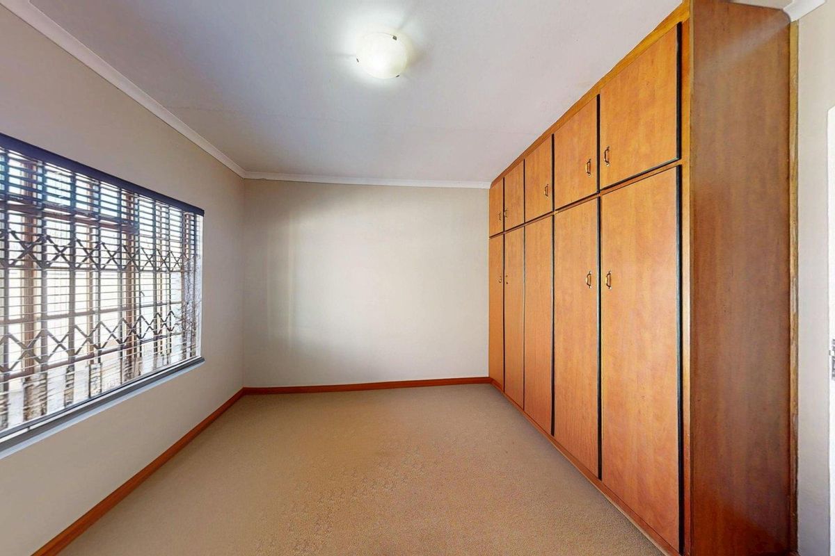 A spacious, empty bedroom featuring wall-to-wall built-in wooden cupboards, carpeted flooring, and a window fitted with security bars.