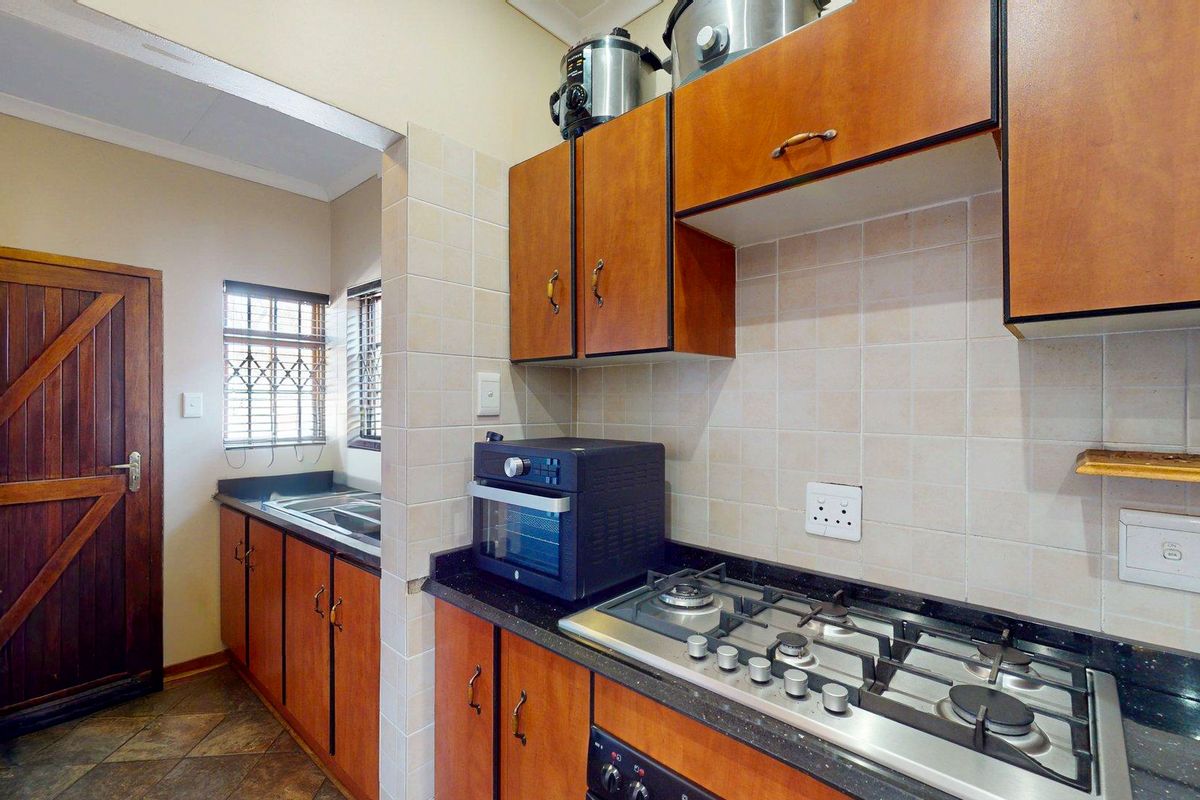 Functional kitchen featuring wooden cabinetry, granite countertops, a gas hob, and a separate scullery area with a sink and security-barred windows.
