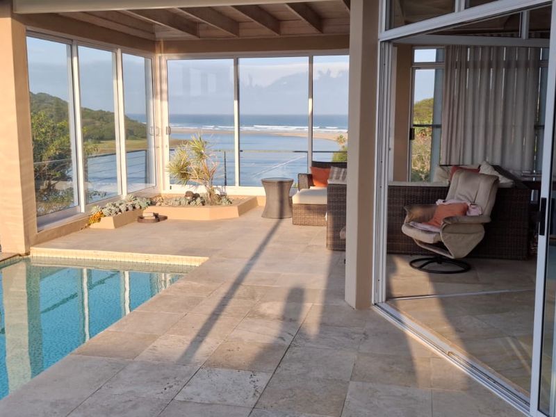 A stunning covered patio area featuring travertine flooring, a private swimming pool, and expansive sliding glass doors offering panoramic coastal and estuary views.