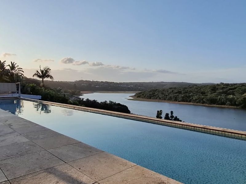 An infinity edge swimming pool overlooking a scenic river and lush landscape, featuring stone patio tiling and a tranquil outdoor setting.