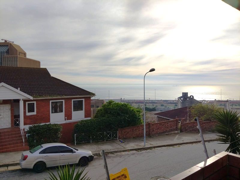 An elevated view overlooking a residential street, brick building, and distant coastal horizon under a cloudy sky.