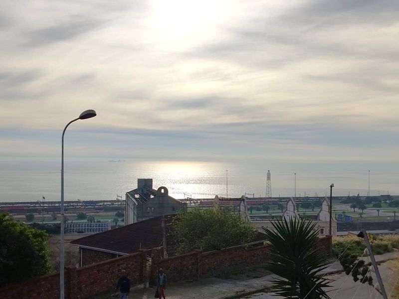 An elevated view overlooking the coastline, industrial rail infrastructure, and surrounding urban landscape under a bright, cloudy sky.