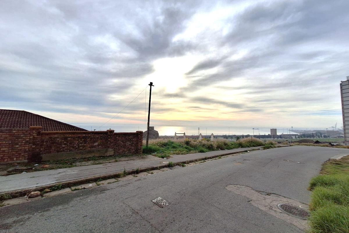 A street-level view showing the approach to the property, featuring a brick boundary wall, asphalt road, and distant coastal horizon.
