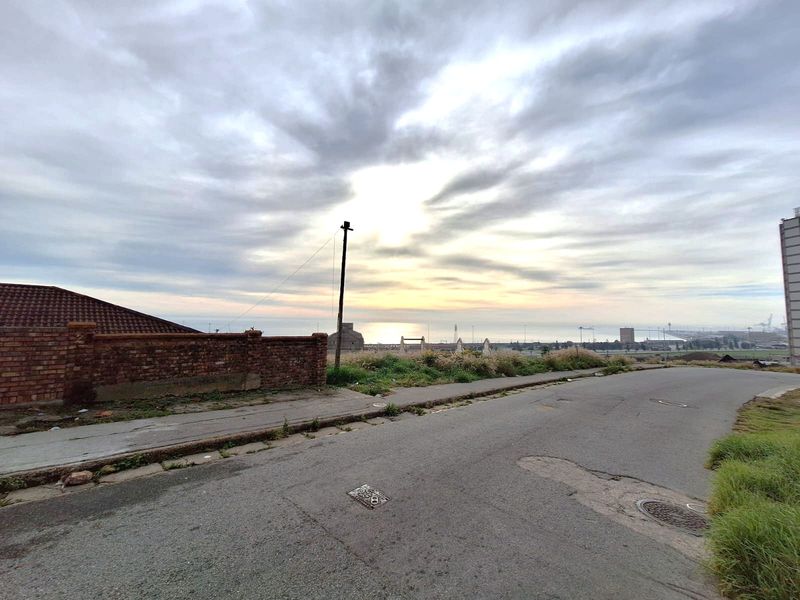 A street-level view showing the approach to the property, featuring a brick boundary wall, asphalt road, and distant coastal horizon.
