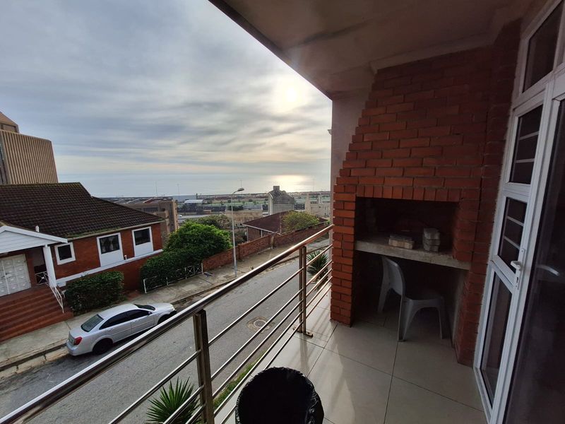 A balcony featuring a built-in brick braai, stainless steel railing, and tiled flooring, offering an elevated view of the surrounding area and coastline.
