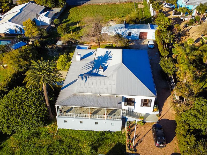 Aerial view of a residential property featuring a metal roof, a spacious covered veranda with railings, and surrounding garden grounds.