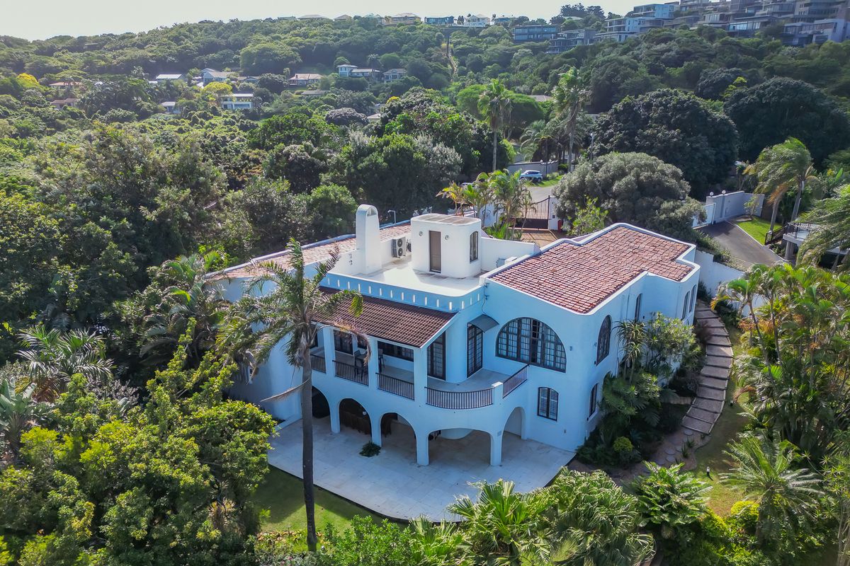 Aerial view of a Mediterranean-style house featuring a terracotta tiled roof, arched veranda, and balcony, set within a lush, landscaped garden.