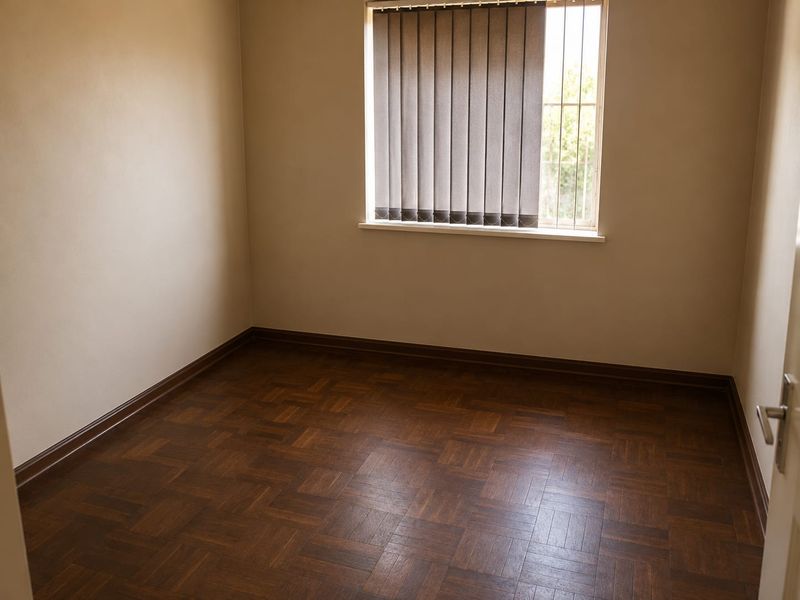 An empty bedroom featuring classic parquet flooring, neutral wall tones, and a window fitted with vertical blinds and security bars.
