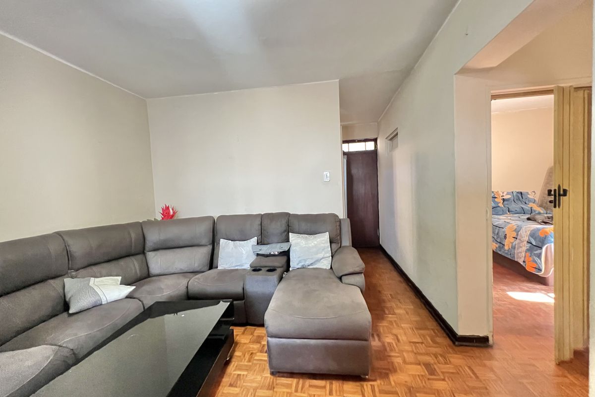 A living room featuring traditional parquet flooring and neutral wall tones, with an open transition leading into an adjacent bedroom area.
