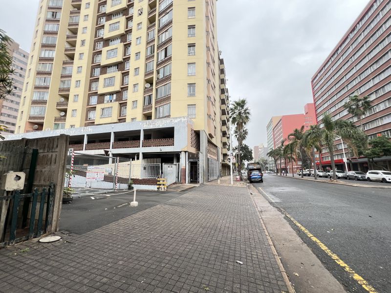Street-level view of a multi-story apartment building featuring a paved sidewalk and adjacent parking structure in an urban setting.