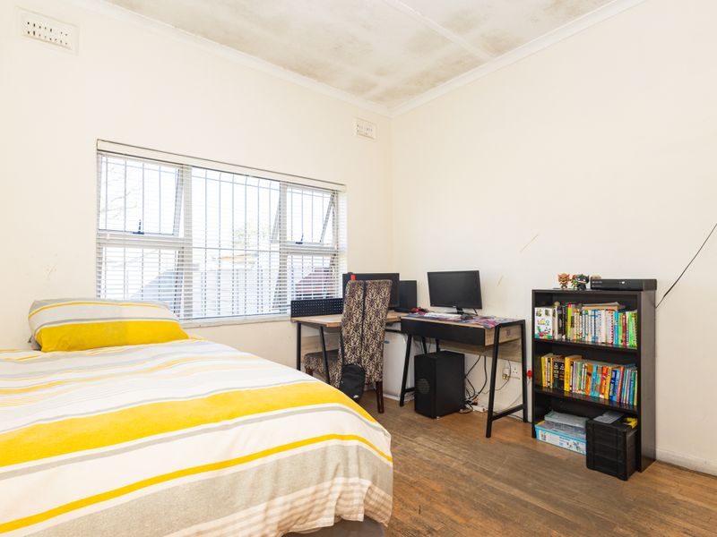 A bedroom featuring timber flooring, window with security bars, and ceiling ventilation. The room shows signs of wear including ceiling staining and scuffed walls.