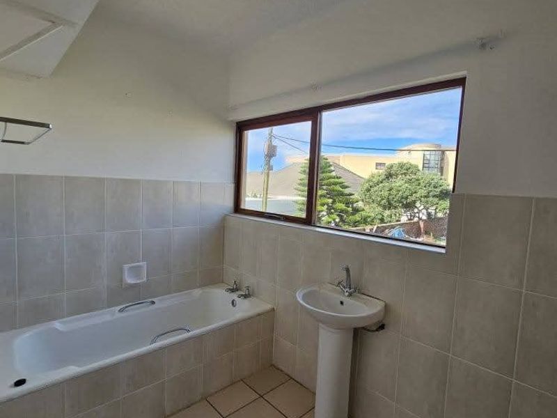 A functional bathroom featuring a built-in bathtub, pedestal basin, and neutral ceramic tiling throughout, offering a clean and simple layout.