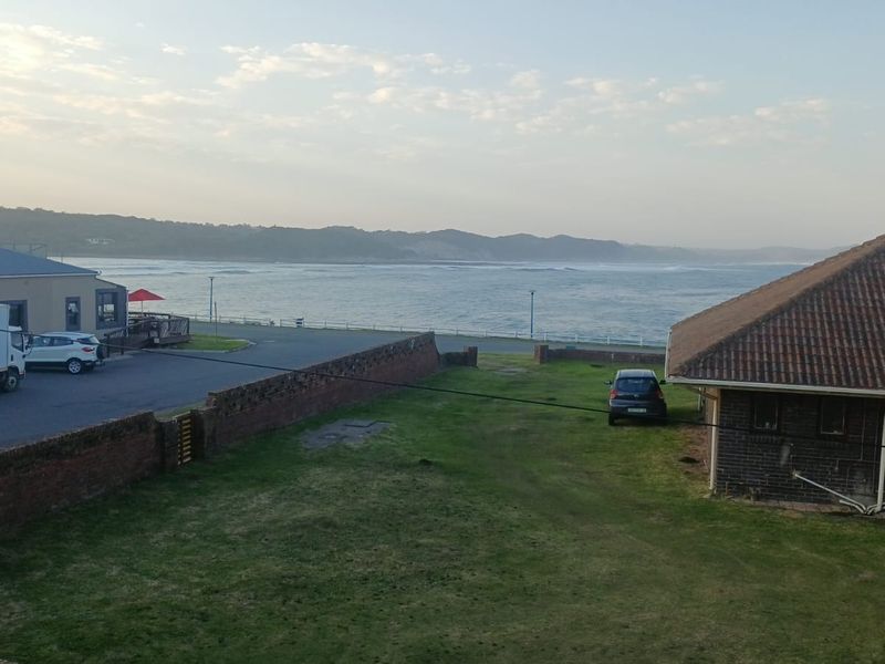 Elevated coastal view overlooking a grassy yard, brick boundary wall, and adjacent buildings with the ocean in the background.