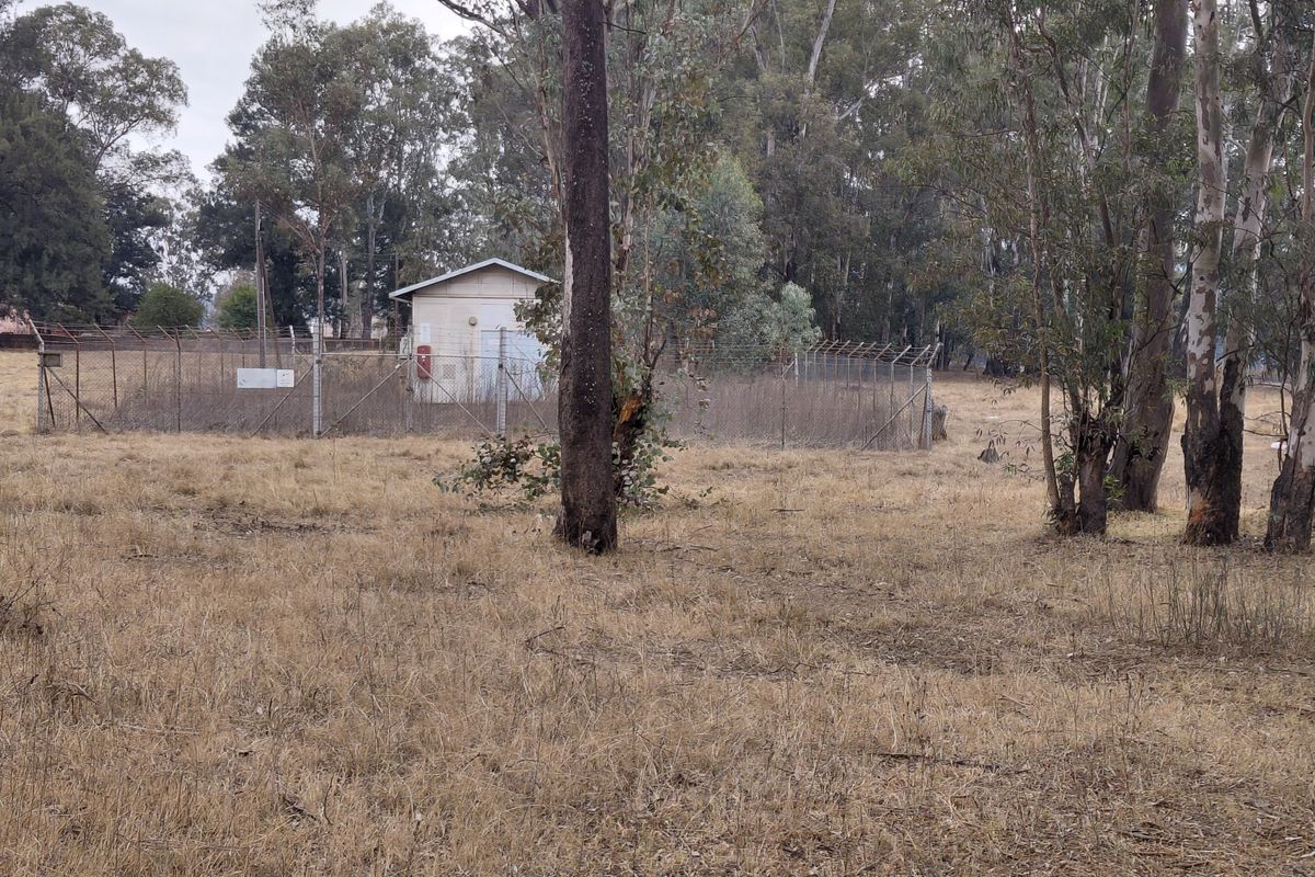 A view of a vacant residential plot featuring a small utility structure enclosed by chain-link fencing and surrounded by mature trees.