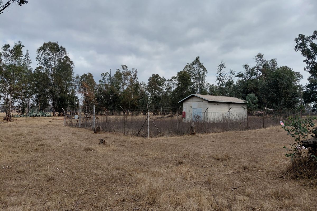 A view of a vacant residential plot featuring a small utility structure enclosed by chain-link fencing amidst dry, overgrown vegetation.
