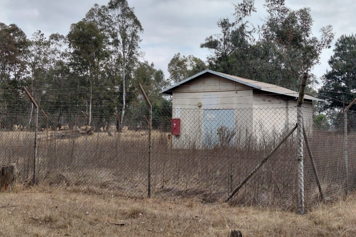 A small utility structure situated on a vacant residential lot, enclosed by chain-link fencing and surrounded by dry, overgrown vegetation.