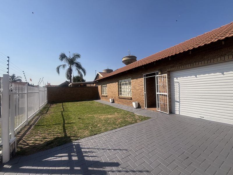 Front exterior view of the townhouse featuring a brick facade, tiled roof, paved driveway, garage, and secure perimeter fencing with a small lawn area.