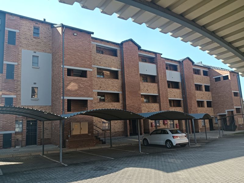 Exterior view of a multi-story brick apartment complex featuring covered parking bays and paved driveway access.