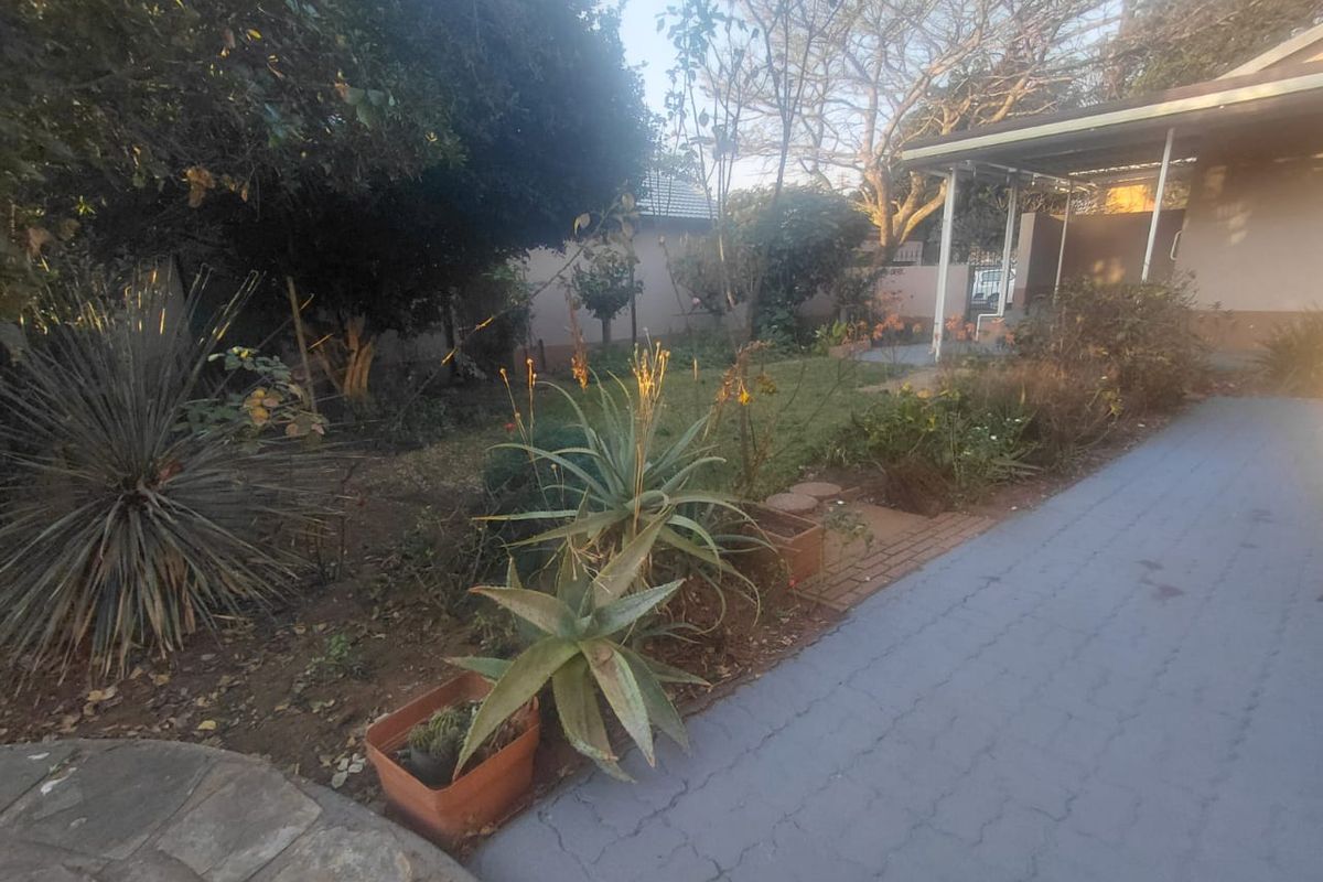 A view of the rear garden area featuring a paved driveway, established landscaping, and a covered patio space.