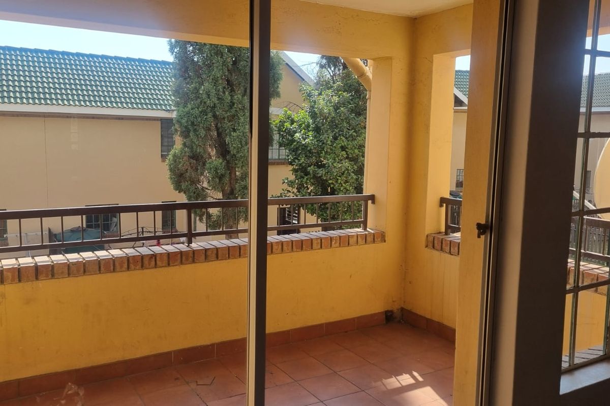 A covered balcony featuring tiled flooring, a brick balustrade with metal railing, and sliding glass door access, offering a view of the surrounding complex.