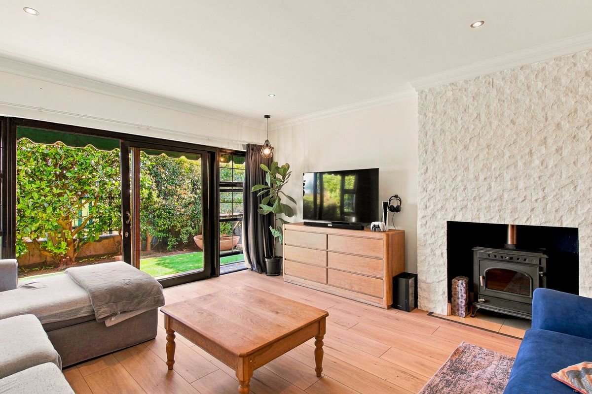 A bright living room featuring timber flooring, a stone-clad wood-burning fireplace, and large sliding glass doors providing direct access to the garden.