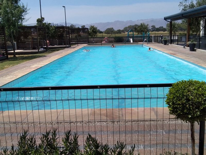 A well-maintained outdoor swimming pool area featuring a paved deck, perimeter fencing, and a timber pergola, set against a scenic mountain backdrop.