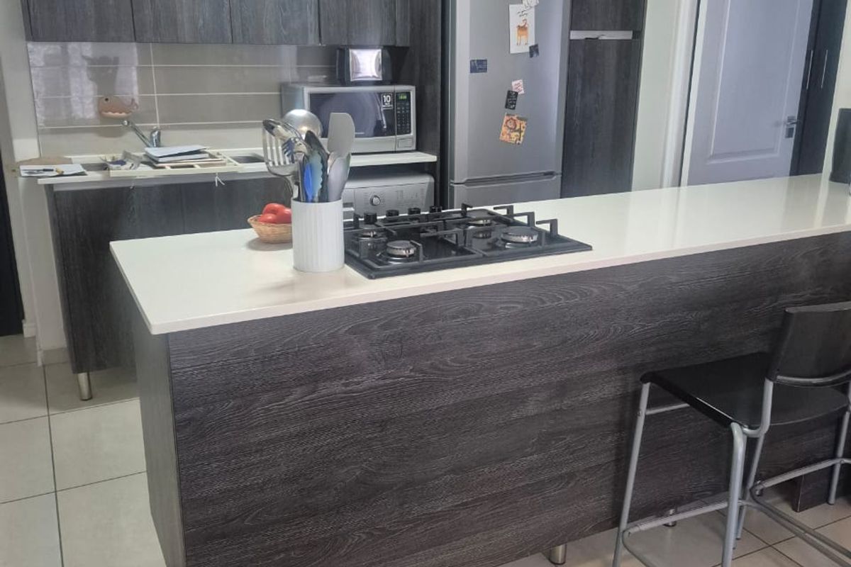 Modern kitchen featuring dark wood-grain cabinetry, a stone-topped breakfast bar with an integrated gas hob, and neutral tiled flooring.