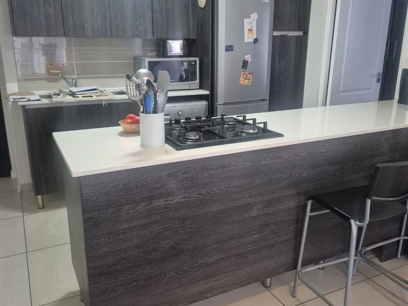 Modern kitchen featuring dark wood-grain cabinetry, a stone-topped breakfast bar with an integrated gas hob, and neutral tiled flooring.