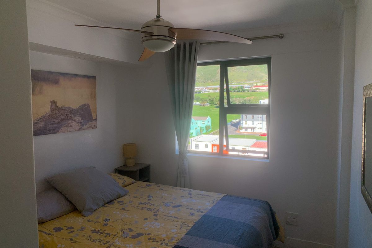 A compact bedroom featuring a ceiling fan, neutral walls, and an aluminium-framed window with views of the surrounding neighborhood.