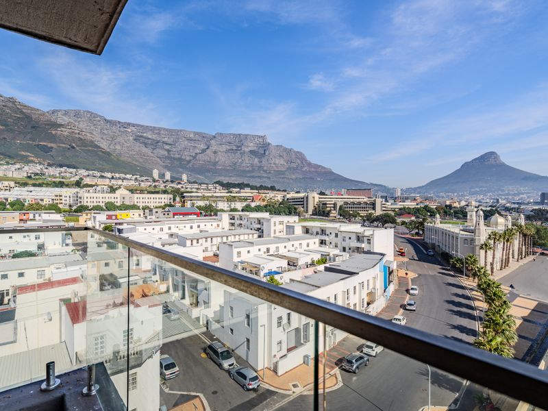 Elevated panoramic view overlooking the city, surrounding buildings, and iconic mountain ranges, captured from a modern apartment balcony with a glass balustrade.