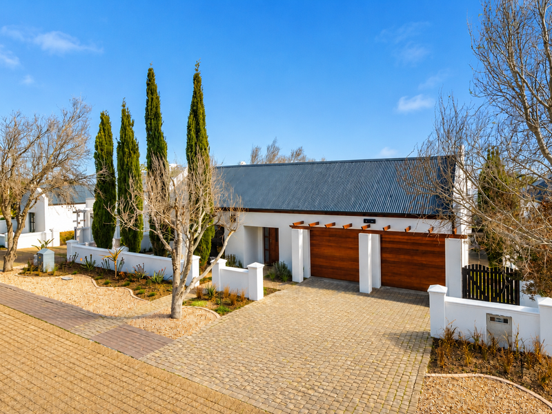 A well-maintained modern home featuring a corrugated metal roof, timber garage doors, a timber pergola, and a neatly paved driveway with landscaped garden beds.