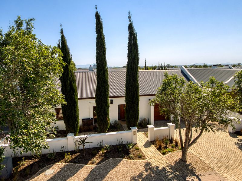 A well-maintained front facade featuring a corrugated metal roof, paved driveway, and landscaped garden with mature cypress trees.