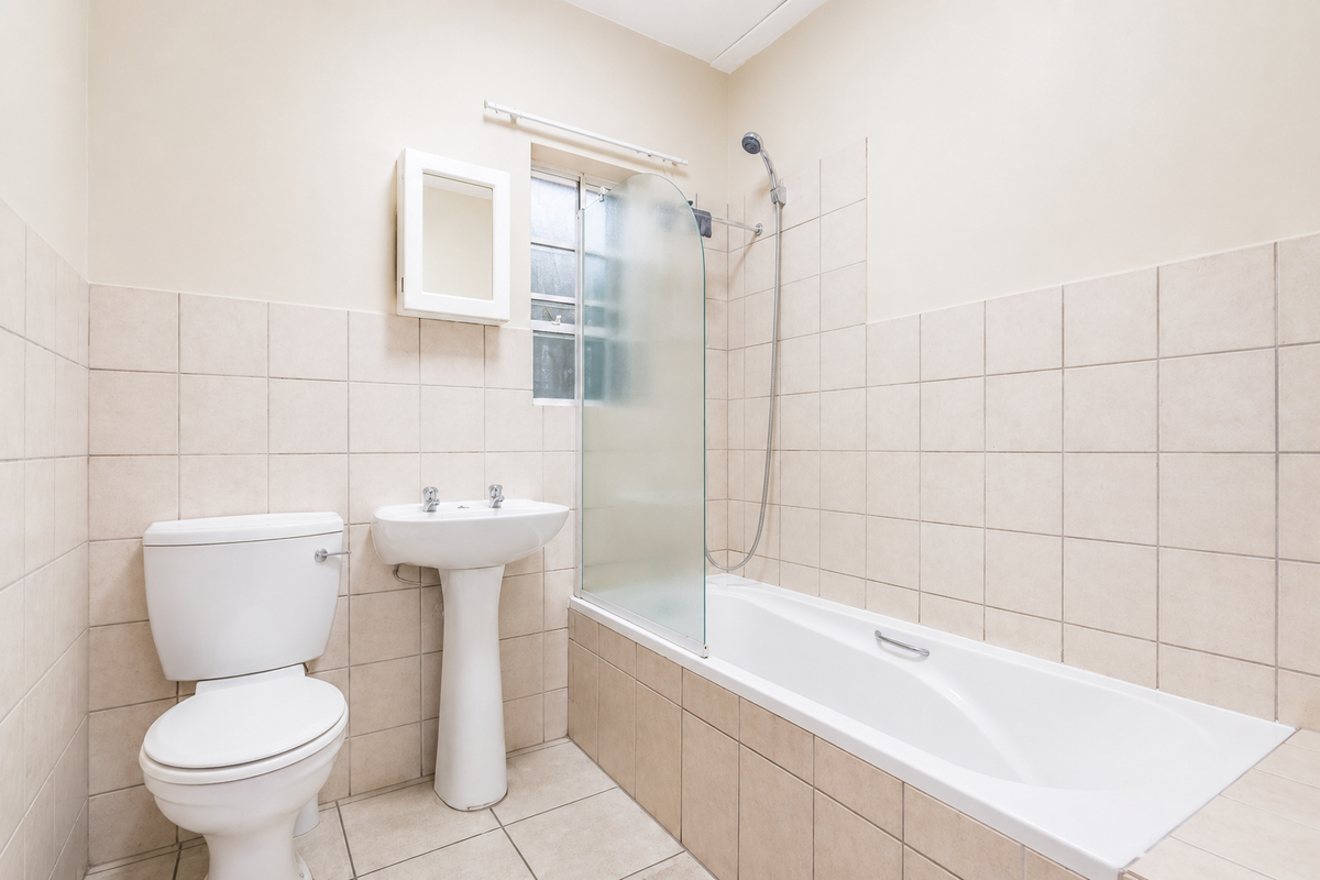 A functional bathroom featuring a pedestal basin, toilet, and a bathtub with a glass shower screen, finished with neutral wall and floor tiling.