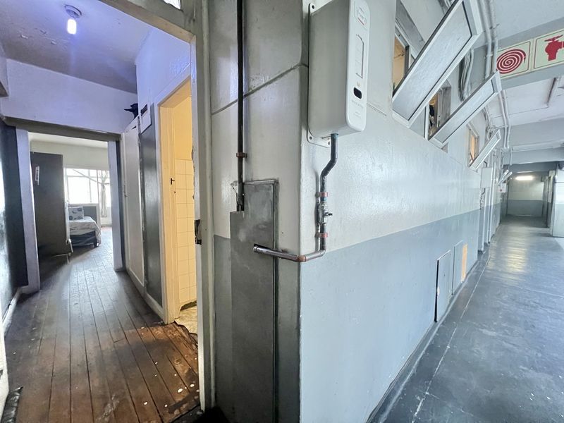 A utilitarian apartment hallway featuring exposed gas geyser plumbing, worn wooden flooring, and access to adjacent rooms.