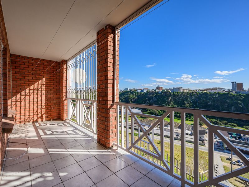A spacious covered balcony featuring exposed brick walls, tiled flooring, and a metal balustrade, offering elevated views of the surrounding area.