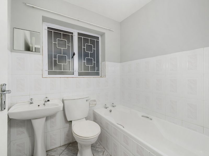 A functional bathroom featuring a pedestal basin, standard toilet, and a bathtub with tiled walls and security bars on the window.
