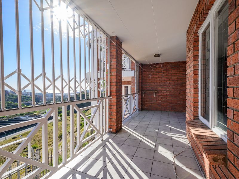 A spacious balcony featuring exposed brick walls, tiled flooring, and secure metal railings, offering elevated views of the surrounding area.